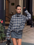 A woman stands outdoors in front of a building, wearing the TIGERMIST LENNEX JACKET in grey check over a plaid turtleneck and short gray skirt. She holds a small black handbag, her hair tied back, with one arm raised near her head.