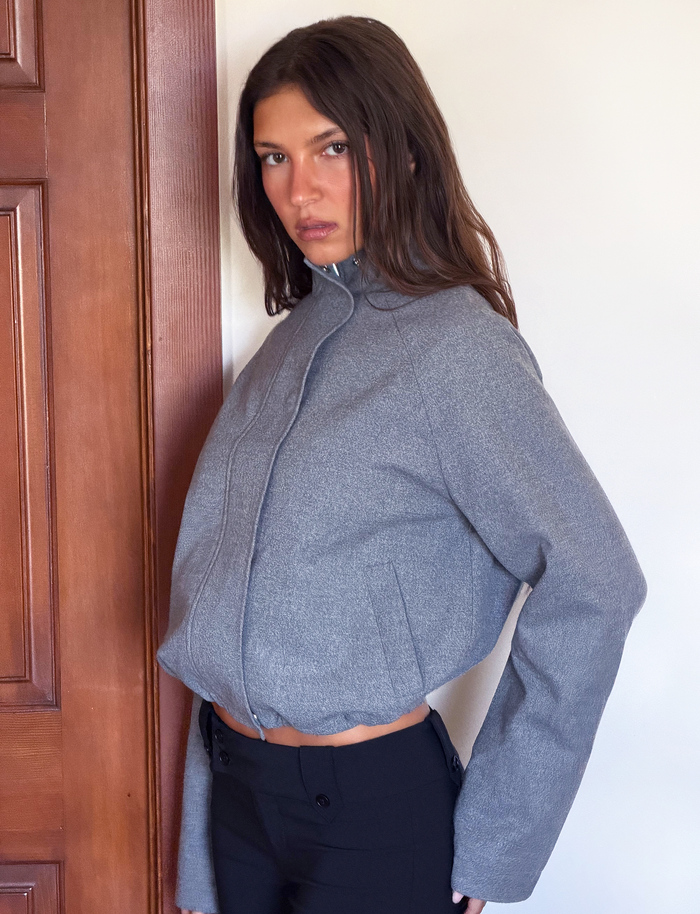 A woman with long brown hair wears the TIGERMIST LENNEX JACKET in heather charcoal, an oversized style with a mock neck collar and zipper deep pockets, paired with black pants. She stands indoors near a wooden door, looking at the camera.