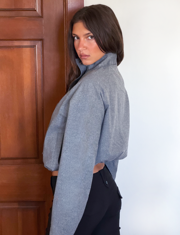 A woman with dark hair glances over her shoulder at the camera, wearing the TIGERMIST LENNEX JACKET in heather charcoal with a mock neck collar, paired with black pants.