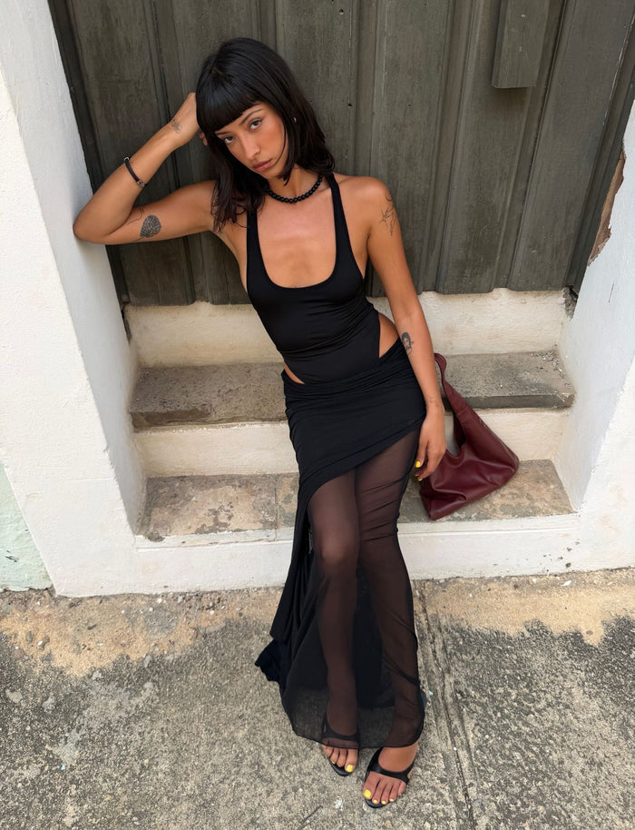 A woman with straight dark hair sits on a stone step wearing the TIGERMIST LILIANA-SUMMER MAXI DRESS in black, paired with black sandals and holding a burgundy bag, as she rests her head on one hand and gazes thoughtfully to the side.