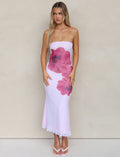 A woman with long blonde hair is wearing the LILLIAN DRESS - PINK by TIGERMIST, a strapless, form-fitting white midi dress featuring large pink floral designs. She poses against a plain beige background and completes the look with white open-toe heels.