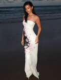 A woman with long, wet hair stands on a beach at dusk, wearing the TIGERMIST LILLIAN MAXI DRESS in Multi Floral Cherry Blossom. Waves and a cloudy sky fill the background.