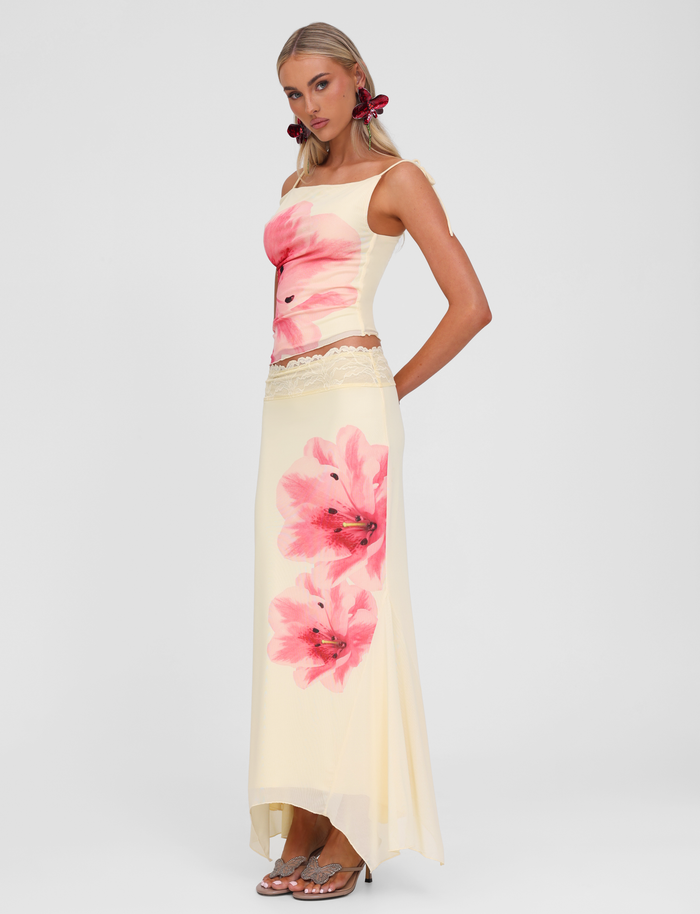 A woman stands in heeled sandals against a plain background, wearing the TIGERMIST LILLIAN SKIRT in yellow with a pink floral print and lace trim waistband, paired with a sleeveless cream top. She has long blond hair and red hair accessories.
