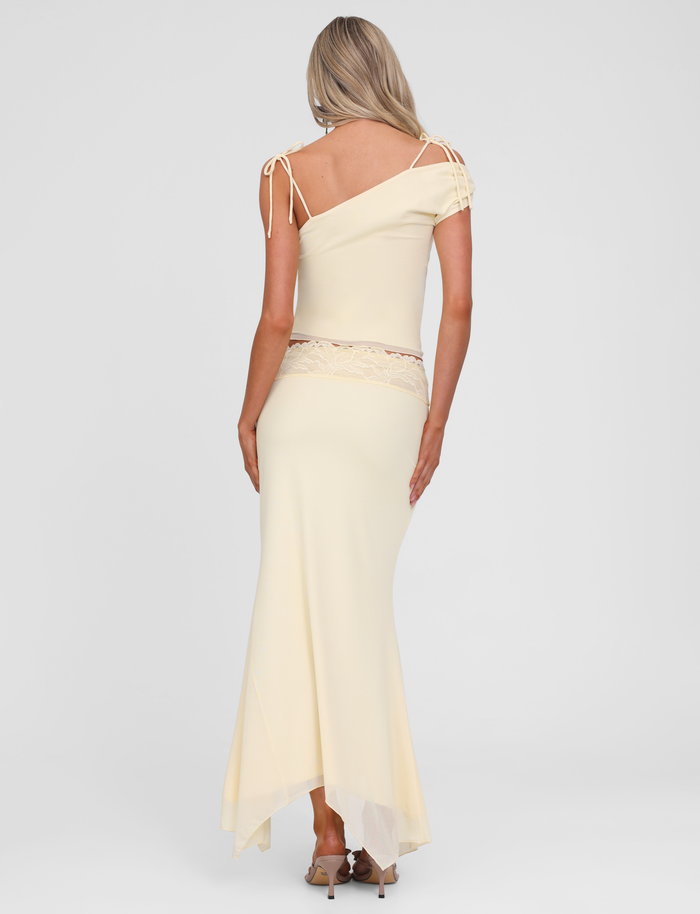 A woman with long blonde hair models the TIGERMIST LILLIAN SKIRT in yellow, detailed with a lace trim waistband and delicate shoulder lace, standing with her back to the camera and wearing high-heeled sandals.