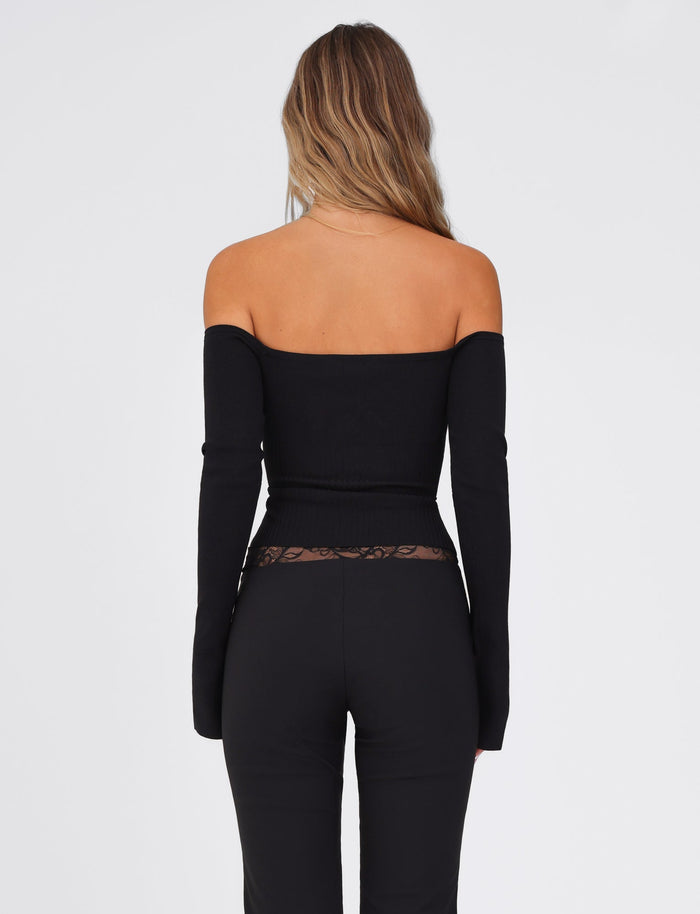 Rear view of a person with long hair, wearing the TIGERMIST LILYANNA LONG SLEEVE TOP in black, paired with high-waisted pants, fully fashioned knit, standing against a plain light background.