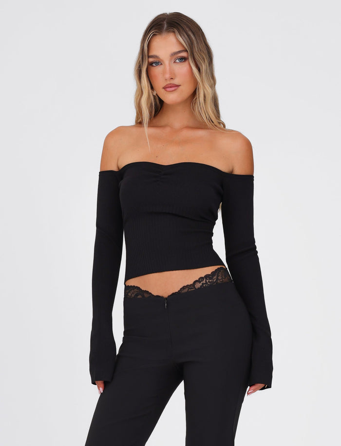 Dressed in the TIGERMIST LILYANNA LONG SLEEVE TOP - BLACK, a person with long hair pairs it with black pants adorned with lace detailing at the waist, standing against a plain white background.