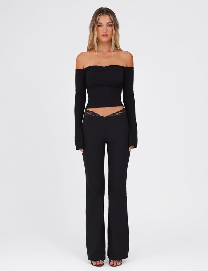 A woman stands against a plain background, showcasing the LILYANA LONG SLEEVE TOP in black from TIGERMIST. This elegant off-the-shoulder, fully fashioned knit is fitted through the body and sleeves, paired with black pants that feature lace detailing. Her hair cascades loosely, and she completes the ensemble with open-toe shoes.