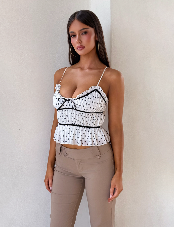 A woman with long brown hair stands against a light wall, wearing the TIGERMIST LIORA TOP - POLKA DOT, a white tiered mesh cami with adjustable straps, paired with beige high-waisted pants. She accessorizes with hoop earrings and has a neutral expression.