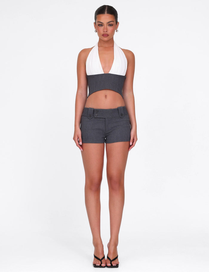A woman faces forward against a white background, wearing the TIGERMIST LOTUS TOP - CREAM : GREY, paired with gray shorts, black heeled sandals, and hoop earrings.