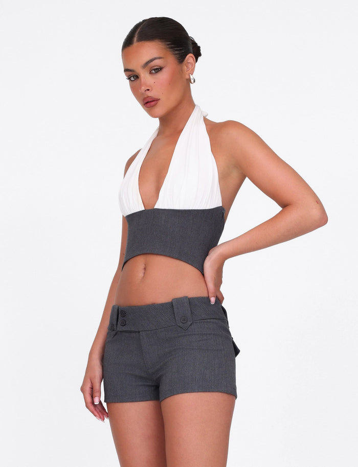 A woman poses confidently in the TIGERMIST LOTUS TOP - CREAM : GREY, a cropped halter made from textured jersey, paired with high-waisted tailored gray shorts with decorative buttons, against a plain white background.