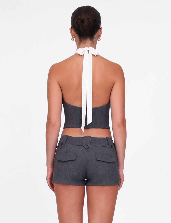 A woman with her hair in a neat bun faces away, wearing the TIGERMIST LOTUS TOP in cream-grey textured jersey and matching high-waisted shorts with a stretch suiting waist and buttoned back pockets, set against a plain white background.