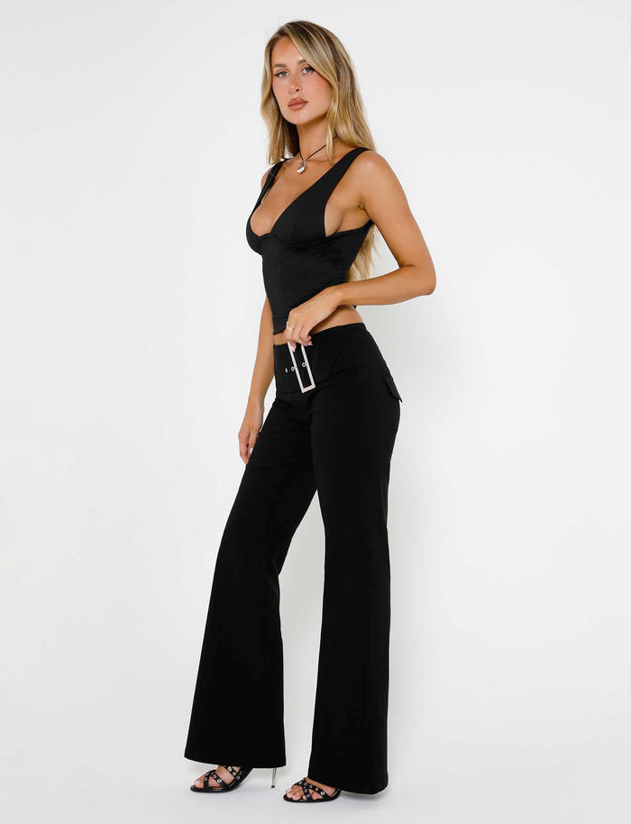 A woman with long blonde hair stands against a plain white background, donning the stylish LUNARIA TOP - BLACK and Tiger Mist black wide-leg pants paired with high-heeled sandals. She holds a small black handbag and gazes slightly to the side.
