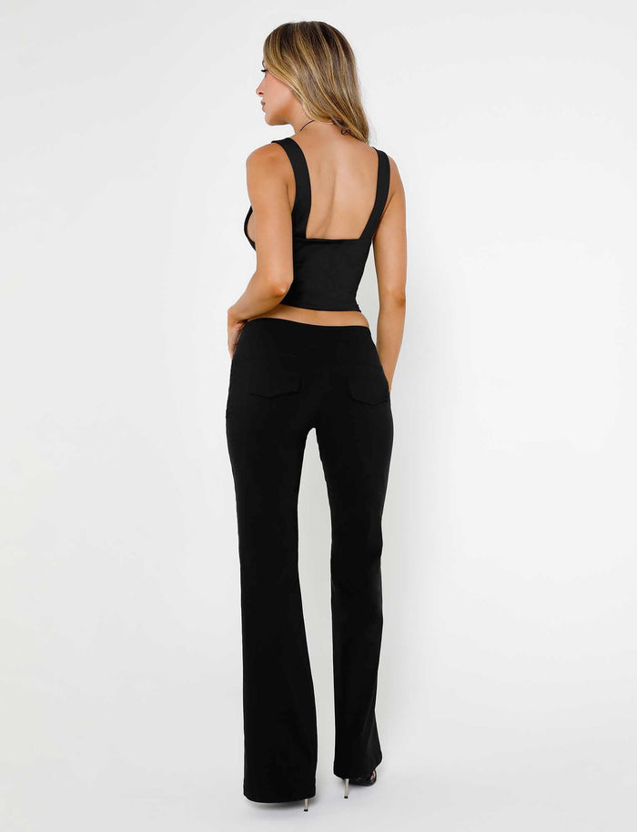 A person with long hair is standing against a plain white background, facing away from the camera. They are dressed in a sleek LUNARIA TOP - BLACK by TIGERMIST with a low back and black high-waisted trousers. They complete the look with high-heeled shoes.