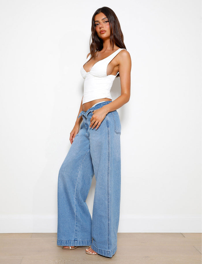 A woman with long, dark hair stands against a white background wearing the LUNARIA TOP - WHITE from TIGERMIST along with light blue wide-leg jeans. She poses with one hand resting on her thigh and the other hanging by her side, complemented by strappy heeled sandals.