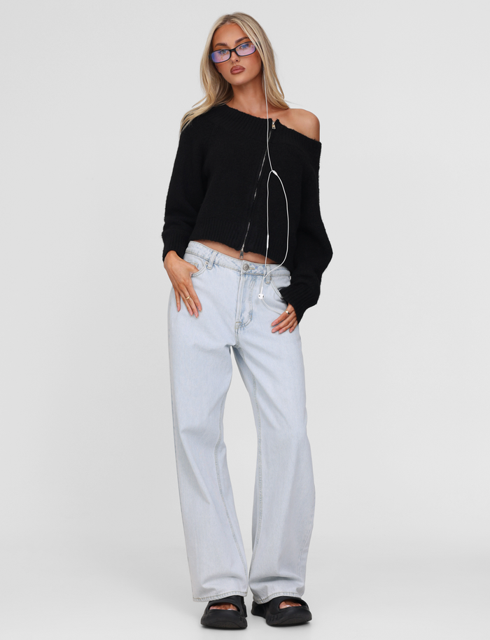 A woman wears the TIGERMIST LUNETTE JUMPER in black, paired with light-wash wide-leg jeans, black sandals, glasses, and earphones around her neck, standing against a plain white background.