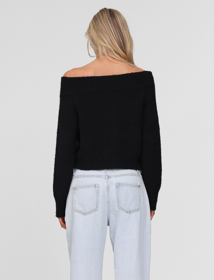 A person with long blonde hair is shown from behind, wearing the TIGERMIST LUNETTE JUMPER in black with light blue high-waisted jeans against a plain light gray background.