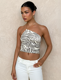 A woman with dark hair in a sleek bun wears the TIGERMIST LYRA TOP - NEUTRAL : ZEBRA, an asymmetric one-shoulder cropped zebra print top with a gold chain strap, paired with white pants, standing against a textured beige wall.