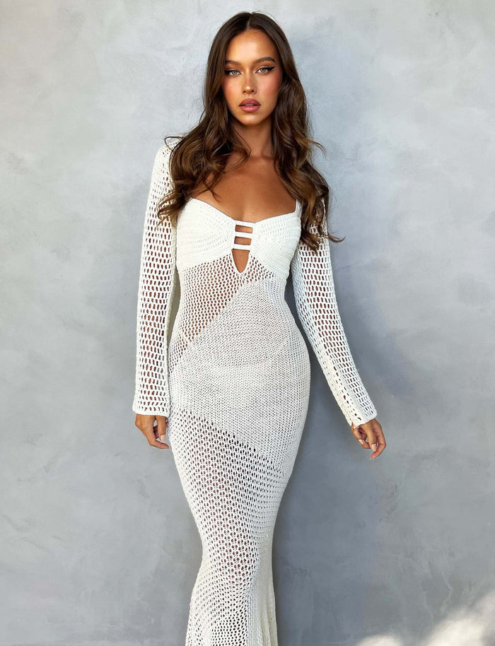 A woman with long wavy hair stands against a gray background, wearing the white Macaulay Dress from TigerMist. The dress features long sleeves, a deep neckline, and cut-out details. She gazes directly at the camera with a serious expression.