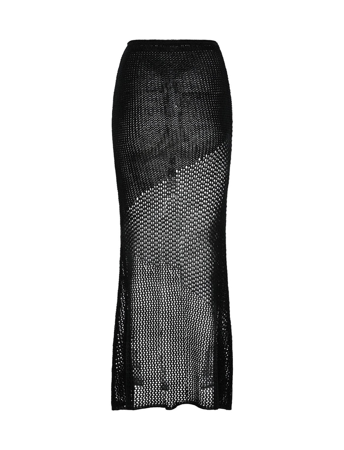 The MACAULEY Skirt in black from TIGERMIST is a full-length mesh skirt featuring a high waistband and a subtle asymmetrical design. The see-through fabric has a textured, open-weave pattern that creates a stylish, sheer effect, showcasing the legs. With its straight hemline and slim fit, it combines elegance with modern flair.