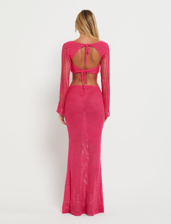A person with long blonde hair facing away from the camera is wearing a bright pink, patchwork crochet two-piece outfit. The top features a unique open-back design with ties, and the bodycon silhouette MACAULEY SKIRT - PINK : HOT PINK from TIGERMIST is floor-length and fits closely to the body. The background is plain white.