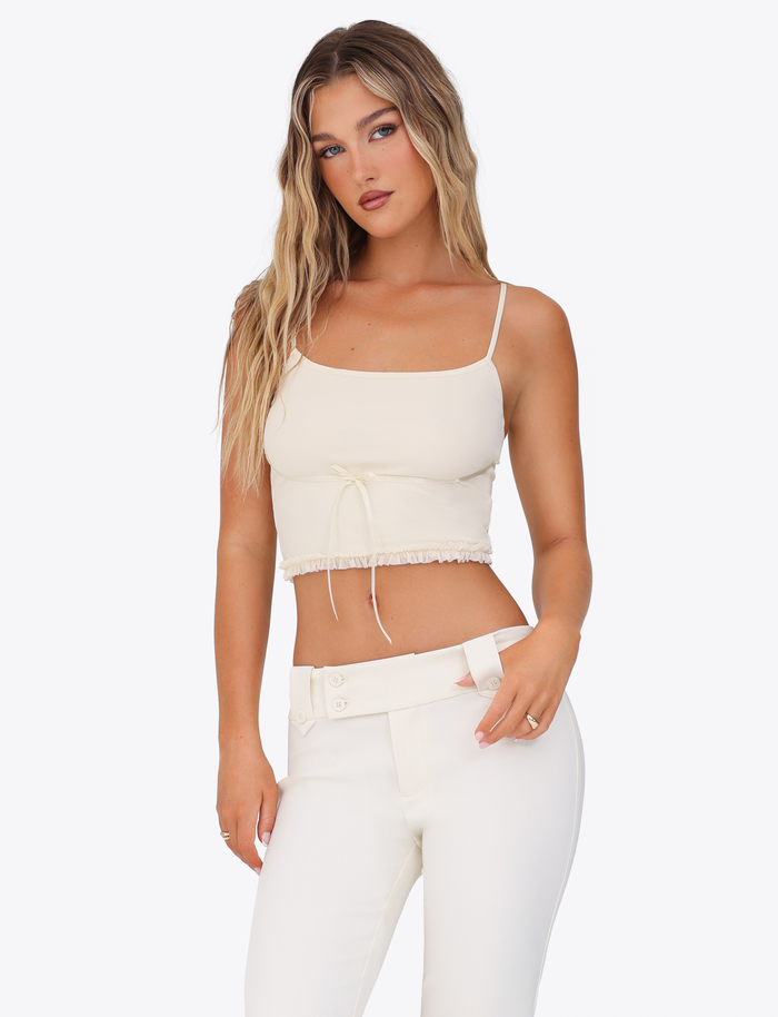 A woman with long wavy blonde hair wears the TIGERMIST MAISIE TOP in pale yellow with adjustable straps and high-waisted white pants, standing against a plain white background, gazing at the camera with a neutral expression.