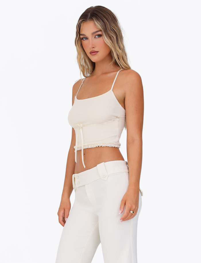 A woman with wavy blonde hair models the TIGERMIST MAISIE TOP in pale yellow, featuring adjustable straps and a tie detail, paired with high-waisted white pants against a plain white background.