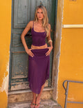 A woman with long blonde hair stands before a weathered green door and yellow wall, wearing a purple sleeveless top and the elegant TIGERMIST MARIANA SKIRT - PURPLE, one hand on her hip and the other on her thigh.
