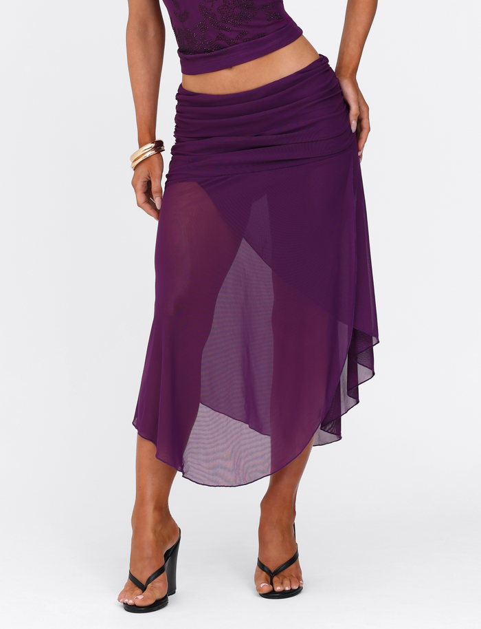 Wearing the TIGERMIST MARIANA SKIRT - PURPLE, a sheer asymmetric wrap skirt with waist ruching, paired with a matching top and black heeled sandals. One hand is on the hip; gold bangles accent the other arm.