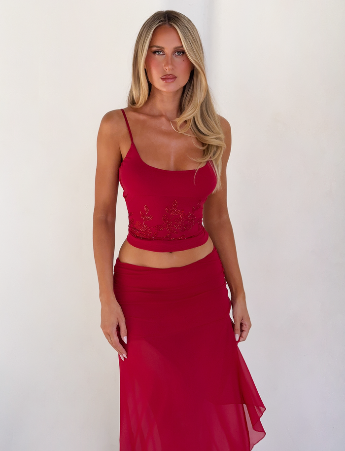 A woman with long blonde hair stands against a white wall, wearing a red sleeveless top and the TIGERMIST MARIANA SKIRT - RED, featuring an asymmetric wrap design. She looks directly at the camera with a neutral expression.
