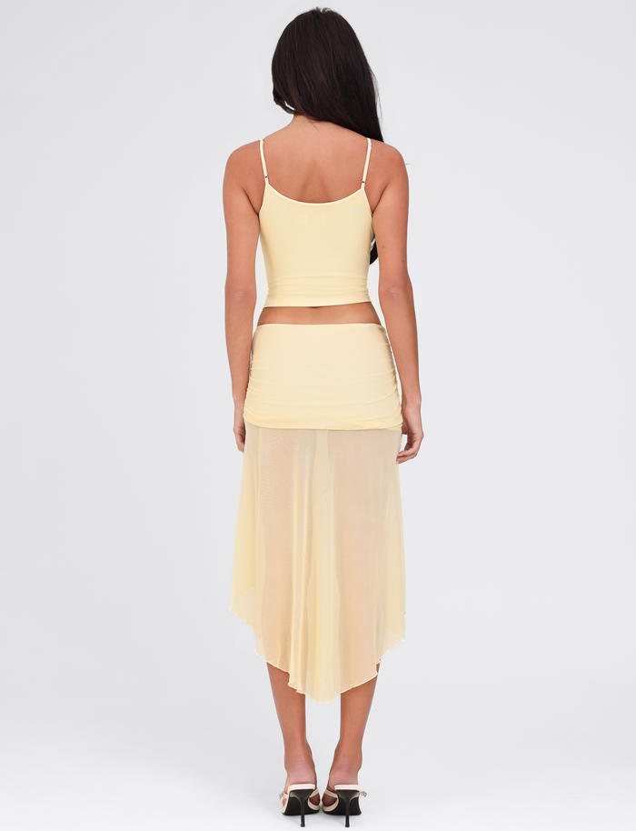 A woman with long dark hair faces away from the camera, wearing the TIGERMIST MARIANA SKIRT in pale yellow and a matching light yellow spaghetti strap top, set against a plain white background.