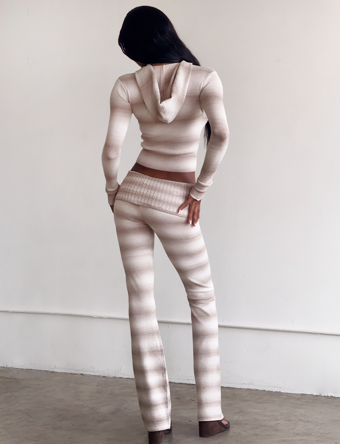 A person with long dark hair faces away, wearing the TIGERMIST MARISSA FOLDOVER KNIT PANT in taupe ombre stripe and a fitted, light beige and white striped hooded top in a minimal indoor space with a plain wall and concrete floor.