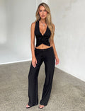 A woman with long blonde hair is standing indoors against a minimalist background. She is wearing a black sleeveless top with a deep neckline and the MARJORIE PANT in black from TIGERMIST. She has a relaxed pose with one hand on her hip and is wearing black sandals.