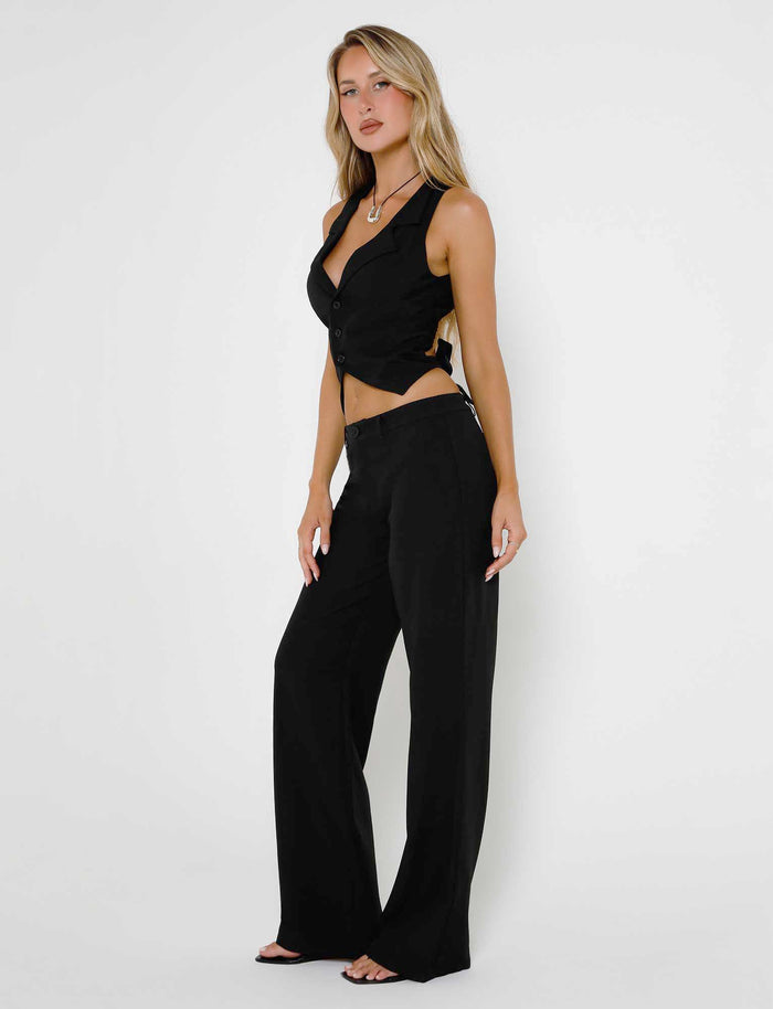 A person with long blonde hair is confidently posing against a plain white background, wearing a stylish black ensemble from TIGERMIST. The outfit consists of the MARJORIE PANT - BLACK, paired with a sleeveless buttoned vest and complemented by black open-toe shoes.