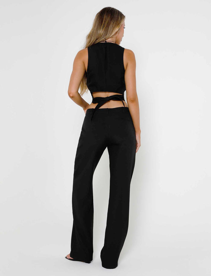 A woman with long, wavy hair stands with her back to the camera, showcasing a TIGERMIST sleeveless black top featuring a halter neck and crisscrossing straps at the back. She pairs it effortlessly with the MARJORIE PANT in black against a plain white background.