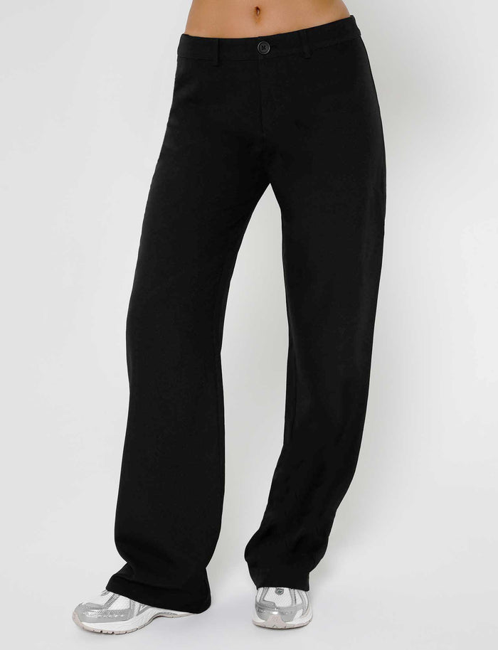 A person is seen standing against a plain background, wearing the MARJORIE PANT - BLACK from TIGERMIST, paired with white sneakers. The pants feature a front button and zipper closure. The person's upper body is not visible in the image.