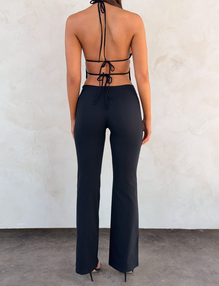 A woman faces a light wall, wearing TIGERMIST’s MARREN PANT in black, styled with a black halter top featuring an open back and tie details, plus black heels. Her arms hang relaxed at her sides.