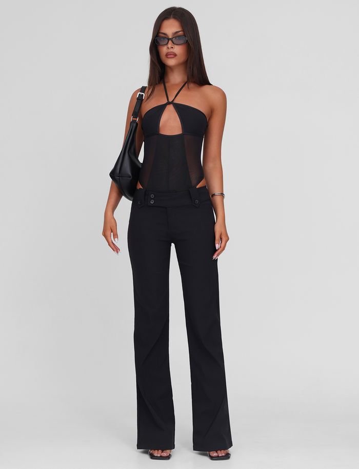A woman poses against a white background wearing the TIGERMIST MELANIA BODYSUIT - BLACK, paired with high-waisted stretch black pants, heeled sandals, dark sunglasses, and a black shoulder bag.
