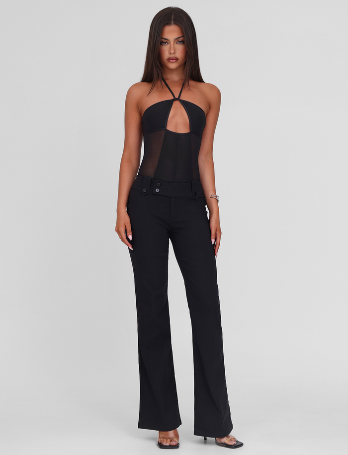 A woman with long straight brown hair wears the TIGERMIST MELANIA BODYSUIT - BLACK, a high-stretch halter neck mesh piece with an adjustable strap, paired with black pants. She stands against a white background, looking at the camera neutrally.