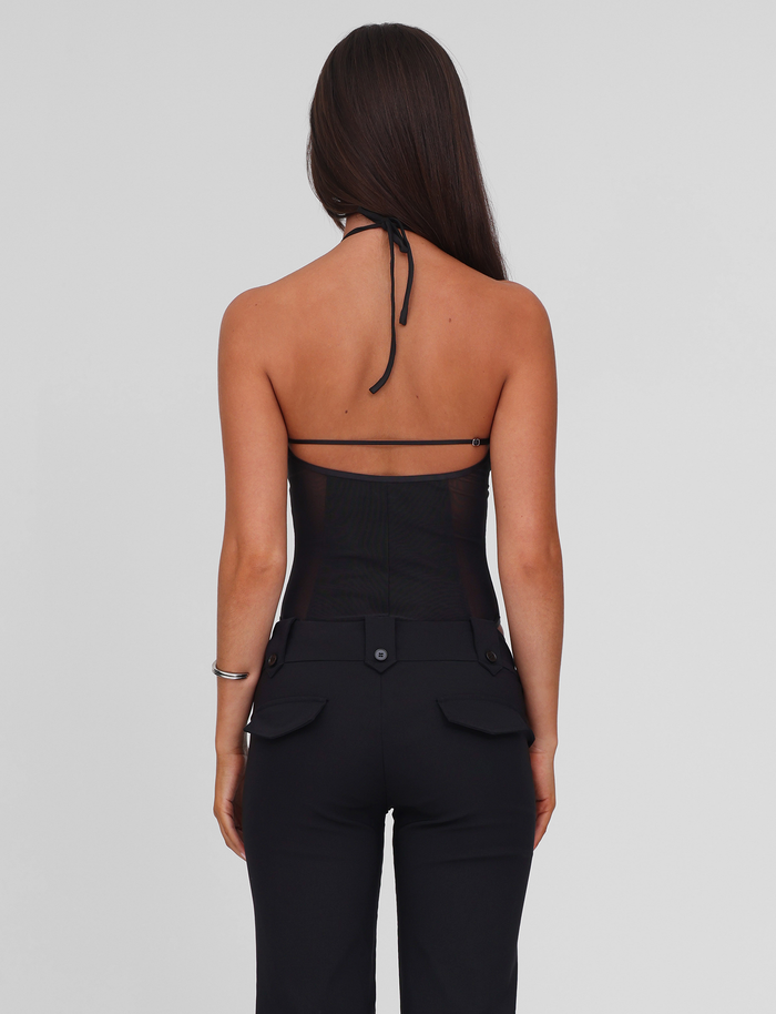 A woman with long brown hair faces away, wearing the TIGERMIST MELANIA BODYSUIT - BLACK with an open back and black pants. Her relaxed pose stands out against a plain light gray background, highlighting the chic style.