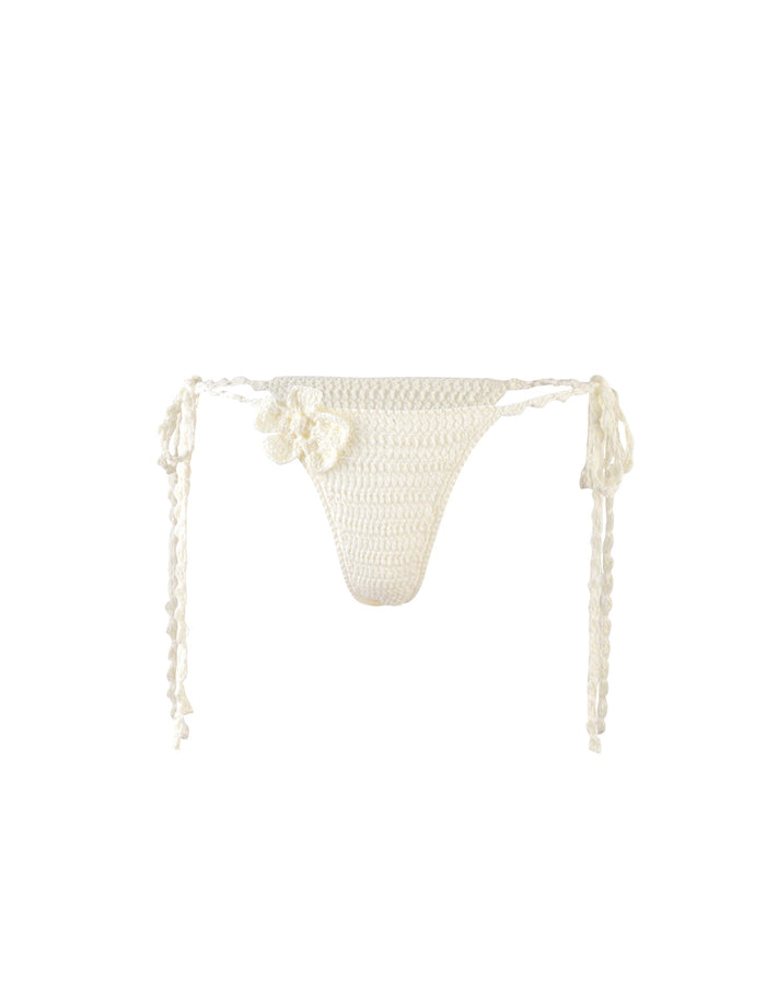 The MELIORA BIKINI BOTTOM - WHITE from TIGERMIST is a white, crocheted bikini bottom featuring side ties and a decorative crocheted flower detail on the left side. The textured, handmade fabric creates a delicate and summery look against the plain white background.