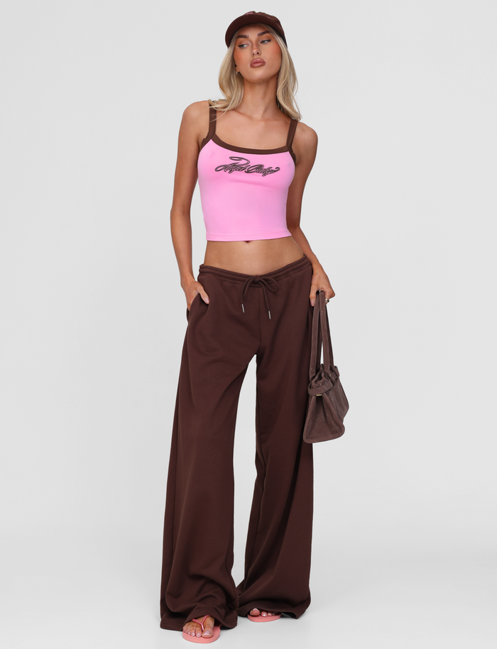 A woman in the TIGERMIST MERRYN TANK - PINK, wide-leg brown pants, a brown cap, pink sandals, and holding a brown handbag stands against a plain white background.