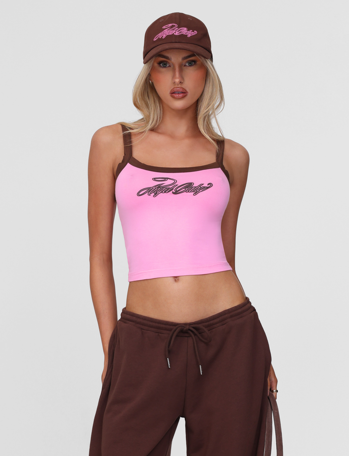 A woman wears the TIGERMIST MERRYN TANK in pink, paired with brown drawstring pants and a brown cap, standing against a plain light background and looking directly at the camera with one hand in her pocket.