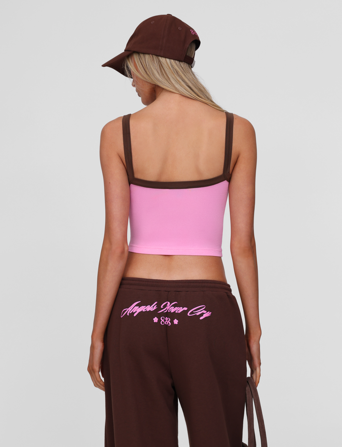 A person with long blonde hair, wearing the TIGERMIST MERRYN CAP - CHOCOLATE, a pink tank top with brown straps, and brown pants with pink lettering on the back, stands with their back to the camera against a plain background.