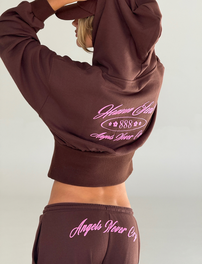 Wearing the TIGERMIST MERRYN TRACK PANT in chocolate, featuring wide legs and pink “Heaven Sent 888” and “Angels Never Cry” embroidery, a person stands sideways with their hood up, face partly hidden.