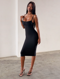 A woman stands indoors against a plain light-colored wall and concrete floor, wearing the TIGERMIST MIDORI BANDAGE MIDI DRESS in black—a fitted, bodycon style—paired with black heels and posing with one hand touching her shoulder.