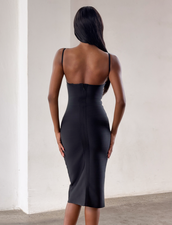 A woman with long dark hair is standing indoors, facing away from the camera, wearing the TIGERMIST MIDORI BANDAGE MIDI DRESS in black, a fitted style featuring thin straps.