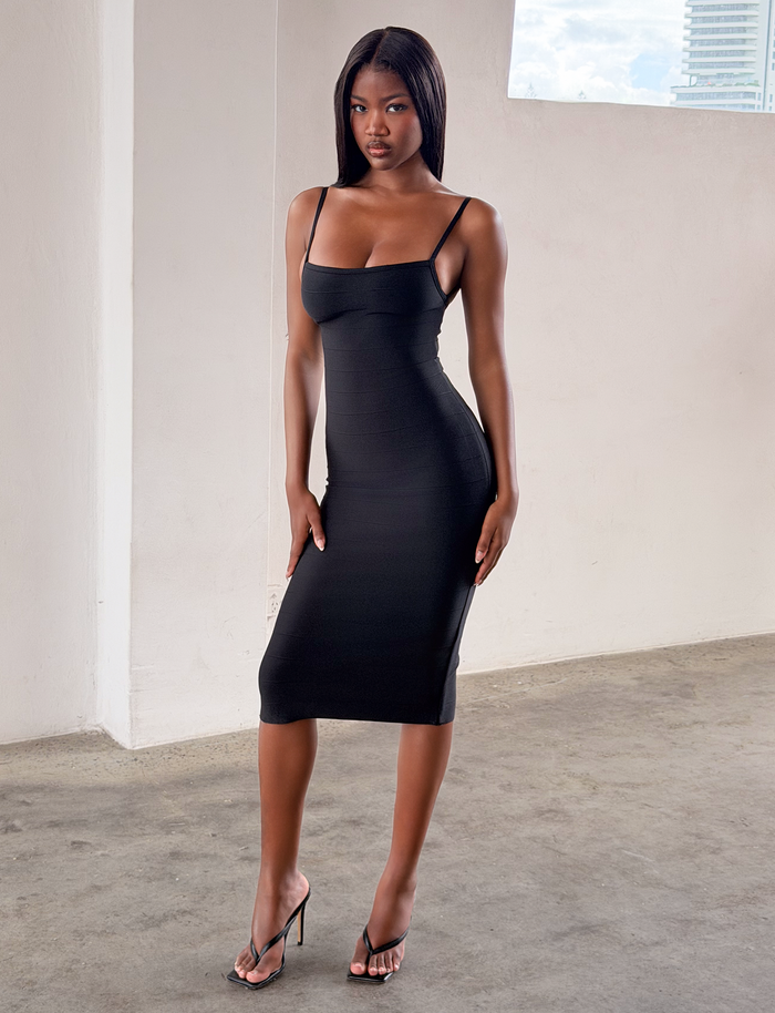 A woman stands confidently indoors near a white wall and window, wearing the TIGERMIST MIDORI BANDAGE MIDI DRESS in black—a fitted, bodycon style—paired with black heels on a concrete floor.
