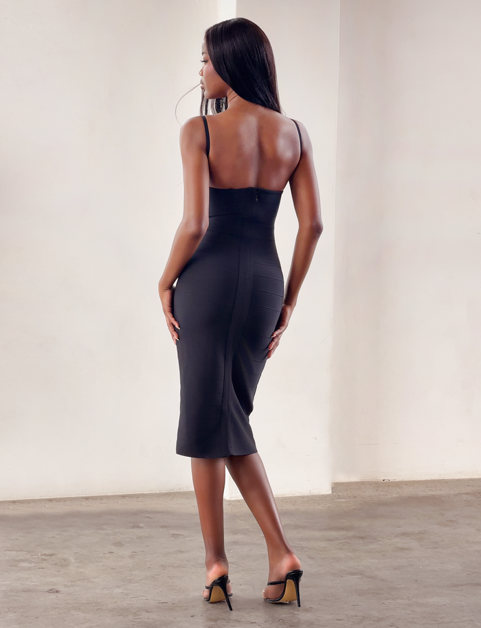 A woman with long hair, wearing the TIGERMIST MIDORI BANDAGE MIDI DRESS in black and black heels, stands on a concrete floor facing away from the camera in a minimalist, light-toned room.