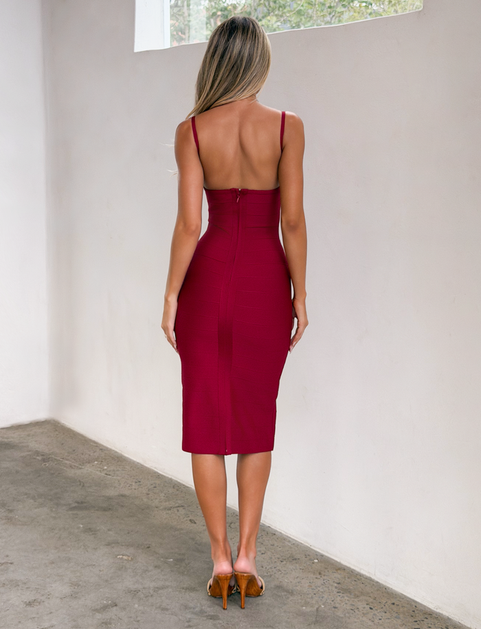 A woman stands indoors facing a white wall, wearing the TIGERMIST MIDORI BANDAGE MIDI DRESS in cherry and tan high heels. Her long hair is down, and she is shown from the back.
