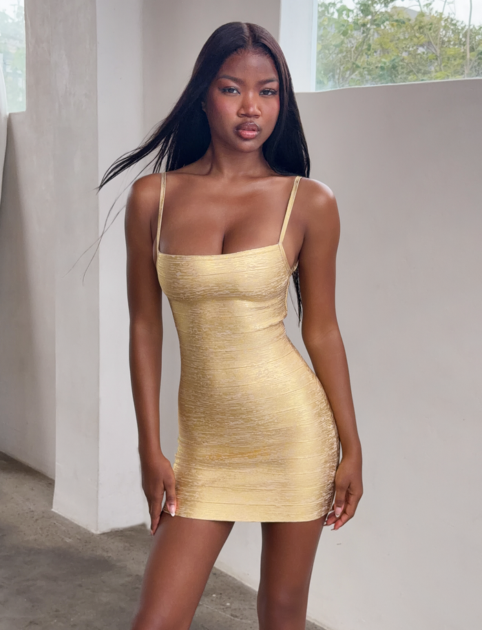 A woman with long straight hair wears the TIGERMIST MIDORI METALLIC BANDAGE MINI DRESS in gold, standing indoors by a window as natural light illuminates the scene.